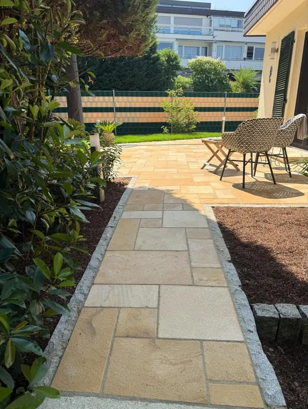 Gartenweg aus sandfarbenen Natursteinplatten mit Granitkante zwischen Beeten mit Rindenmulch, der zu einer Terrasse aus demselben Stein führt